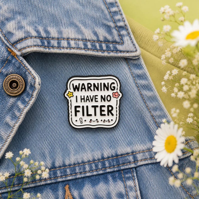 Warning I Have No Filter hard enamel pin displayed on a light blue denim jacket against a soft sage green background with daisy flowers. Funny teacher pin badge, coworker gift, office humour accessory, jacket pin, backpack badge and unique enamel gift by Pocketing Positivity.