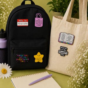 Funny mental health enamel pin badges displayed on a black backpack and cream canvas tote bag. Features Hello I'm Dying Inside, Not Everything That Weighs You Down Is Yours to Carry, Hang On Let Me Overthink This, You Didn't Totally Suck Today, In a Really Good Place Right Now Not Mentally book pin, and Hold On Let Me ChatGPT This enamel badge. Perfect gift for teachers, coworkers, students, office lanyards, backpacks and tote bags.