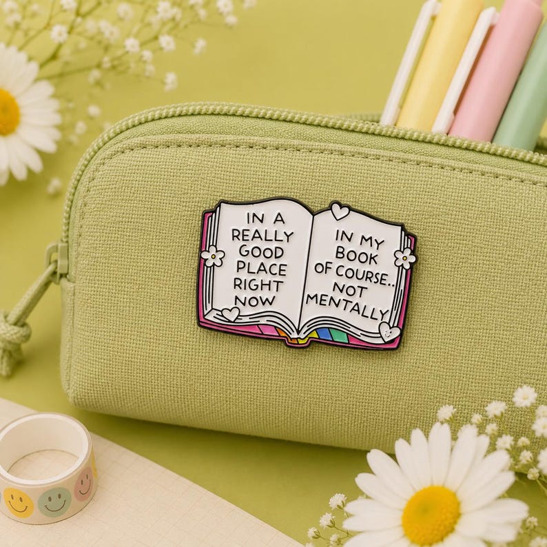 Open book hard enamel pin attached to a sage green pencil case surrounded by pastel pens and daisy flowers. Funny book lover enamel badge reading "In a really good place right now... in my book of course, not mentally." Perfect teacher gift, librarian gift, reader accessory, backpack pin and stationery lover gift.