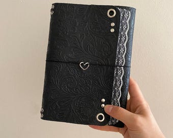 May include: A black journal cover featuring an embossed floral pattern, white lace trim along the right edge, silver metal grommets, and a black elastic band closure secured with a silver heart-shaped charm centered on the front cover.