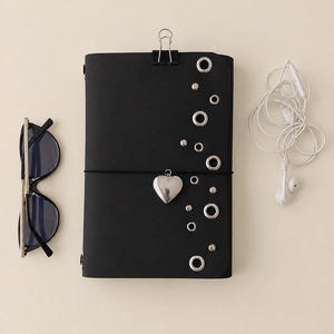 May include: A black notebook with a silver heart charm, metal eyelets, and a binder clip at the top. To the left are black-framed sunglasses, and to the right are white wired earbuds. The notebook is secured with a thin black elastic band.