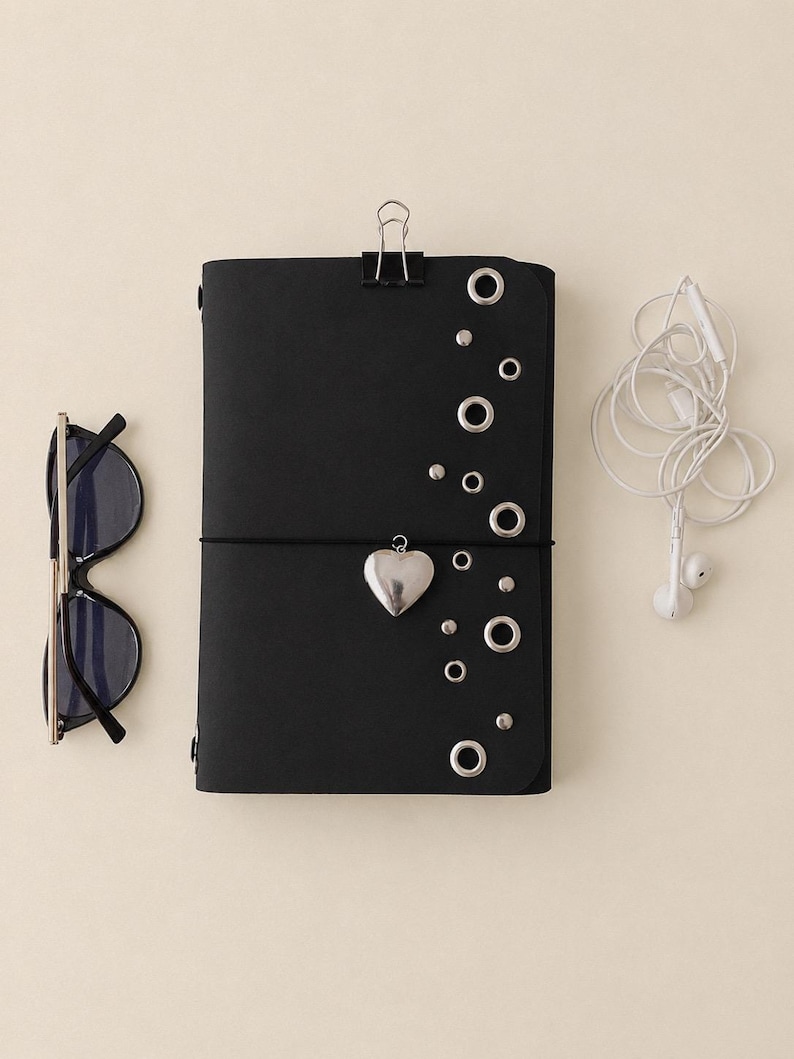 May include: A black notebook with a silver heart charm, metal eyelets, and a binder clip at the top. To the left are black-framed sunglasses, and to the right are white wired earbuds. The notebook is secured with a thin black elastic band.