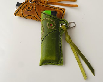 May include: Two leather lighter cases, one in mustard yellow and one in lime green, each featuring black stitched swirl patterns, a metal eyelet, and a matching leather cord tie. The green case holds a green lighter, while the yellow case holds a black lighter.