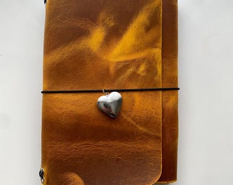 May include: A rectangular notebook with a distressed golden-brown leather cover. A thin black elastic band wraps horizontally around the center, securing a polished silver-toned heart-shaped charm that hangs from the front of the cover.