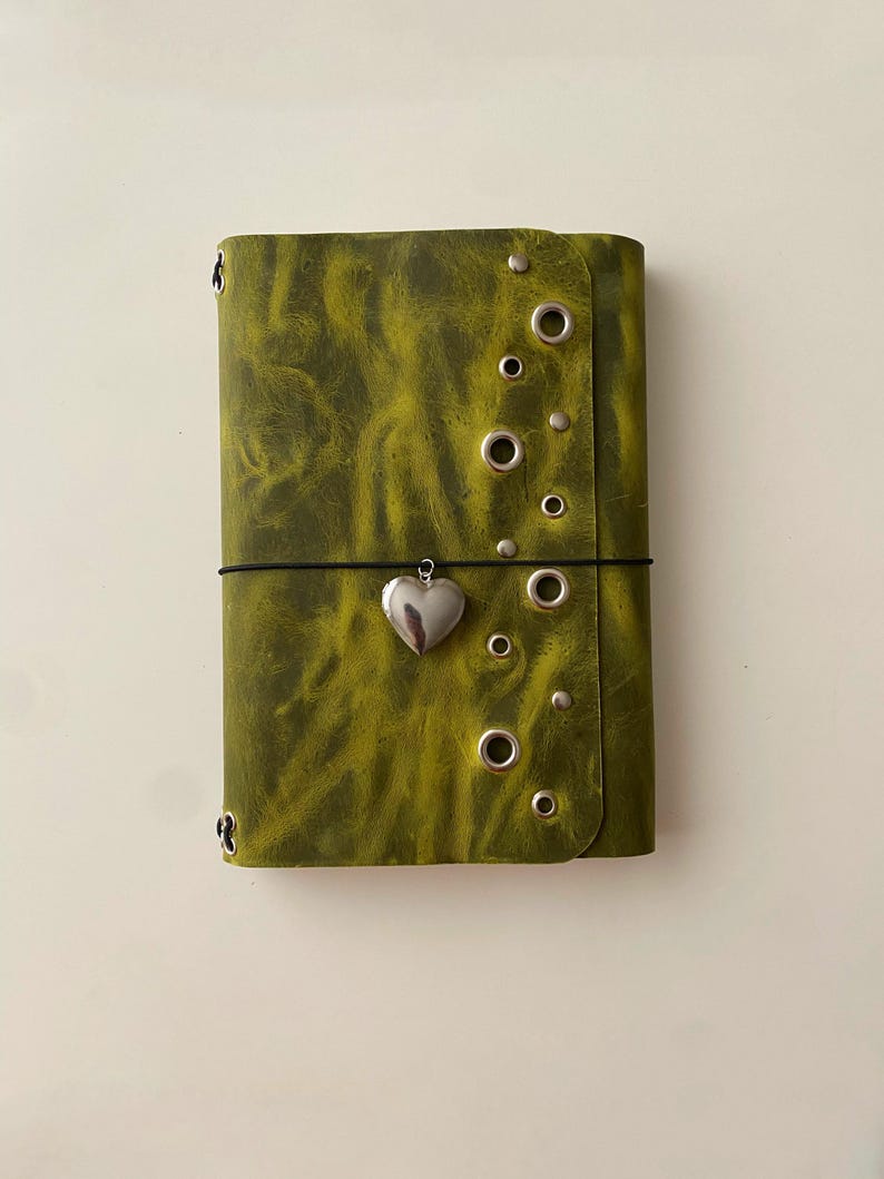 May include: A rectangular olive green journal with a marbled texture, featuring a silver heart-shaped charm attached to a black elastic band closure. The front cover is decorated with several silver-toned metal grommets and small studs.