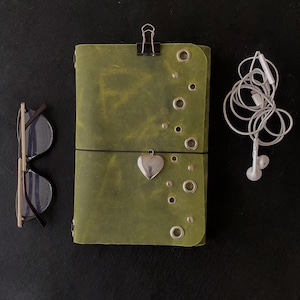 May include: A rectangular olive green leather journal featuring silver-toned metal grommets and studs, a black binder clip at the top, and a silver heart-shaped charm attached to a black elastic band. Beside it are black-framed glasses and white wired earbuds.