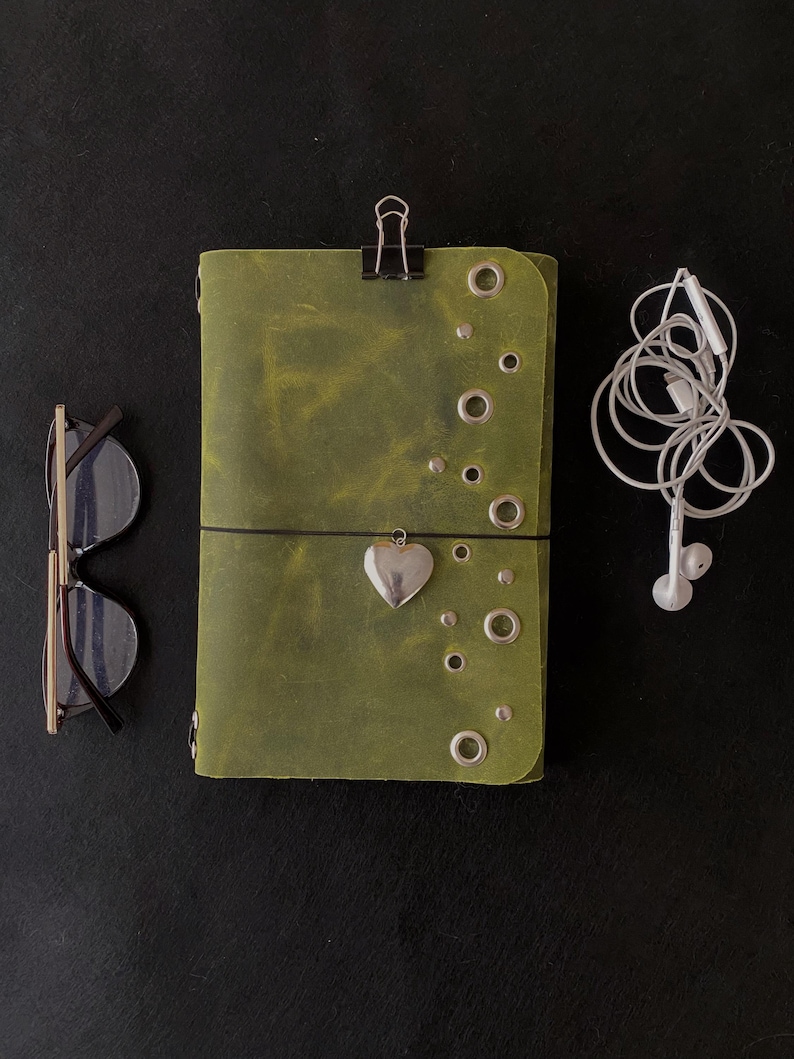 May include: A rectangular olive green leather journal featuring silver-toned metal grommets and studs, a black binder clip at the top, and a silver heart-shaped charm attached to a black elastic band. Beside it are black-framed glasses and white wired earbuds.