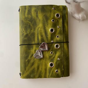 May include: A rectangular olive green journal with a textured finish, featuring a vertical row of silver metal grommets and studs along the right edge. A thin black elastic band wraps around the cover, holding a small silver heart-shaped locket charm.