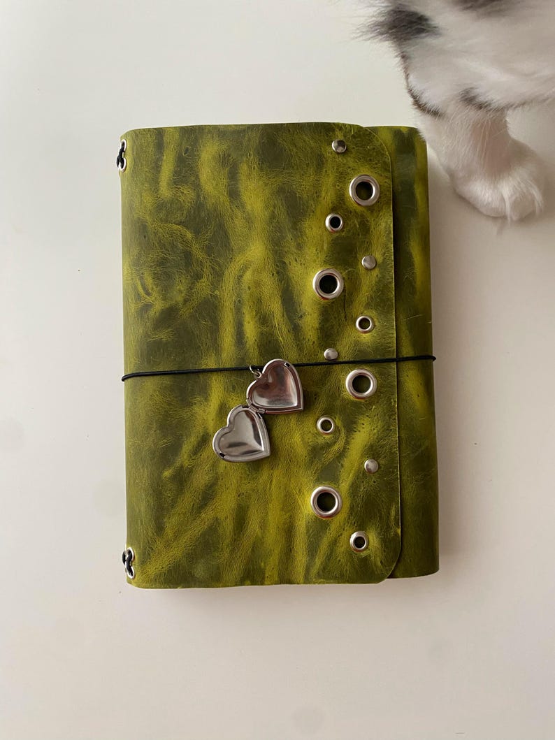 May include: A rectangular olive green journal with a textured finish, featuring a vertical row of silver metal grommets and studs along the right edge. A thin black elastic band wraps around the cover, holding a small silver heart-shaped locket charm.