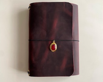 May include: A dark burgundy leather journal with a smooth, matte finish. A thin black elastic cord wraps horizontally around the center, securing a gold-toned oval charm featuring a smooth, vibrant red stone inlay at the front.