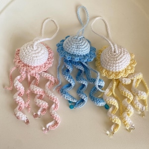 Puede incluir: Tres adornos de medusas de ganchillo hechos a mano en colores pastel. Cada medusa tiene una parte superior blanca, un borde con volantes y largos tentáculos rizados. Uno es rosa, uno es azul y uno es amarillo, con pequeñas cuentas y dijes.