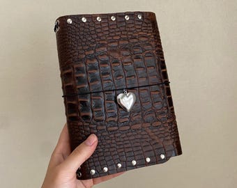 May include: A dark brown journal with a textured crocodile skin pattern, featuring a row of silver-toned metal studs along the top and bottom edges. A thin black elastic band wraps around the center, holding a polished silver heart-shaped charm.