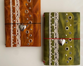 May include: Two rectangular journals with textured covers in tan and olive green. Each features a vertical strip of white lace, silver-toned metal studs, and grommets. A silver heart-shaped charm hangs from a thin elastic band on each cover.