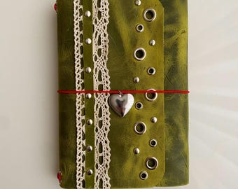 May include: A green leather journal featuring a vertical strip of white lace trim, silver metal studs, and a series of silver grommets. A thin red elastic cord wraps horizontally around the cover, holding a silver heart-shaped charm.