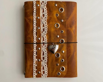May include: A tan leather journal featuring a vertical strip of white lace trim secured by silver metal studs. The cover is decorated with scattered silver metal grommets and a central black elastic band holding a silver heart-shaped charm.
