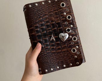 May include: A dark brown journal cover with a textured crocodile skin pattern, decorated with silver-toned metal studs and grommets along the edges. A black elastic band wraps around the center, holding a silver letter A charm and a silver heart charm.