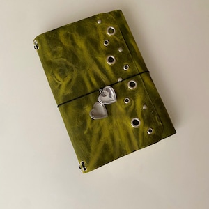 May include: A rectangular olive green journal cover with a distressed texture, featuring a vertical row of silver metal eyelets and studs along the right side. A thin black elastic band wraps around the center, holding a small silver heart-shaped locket.