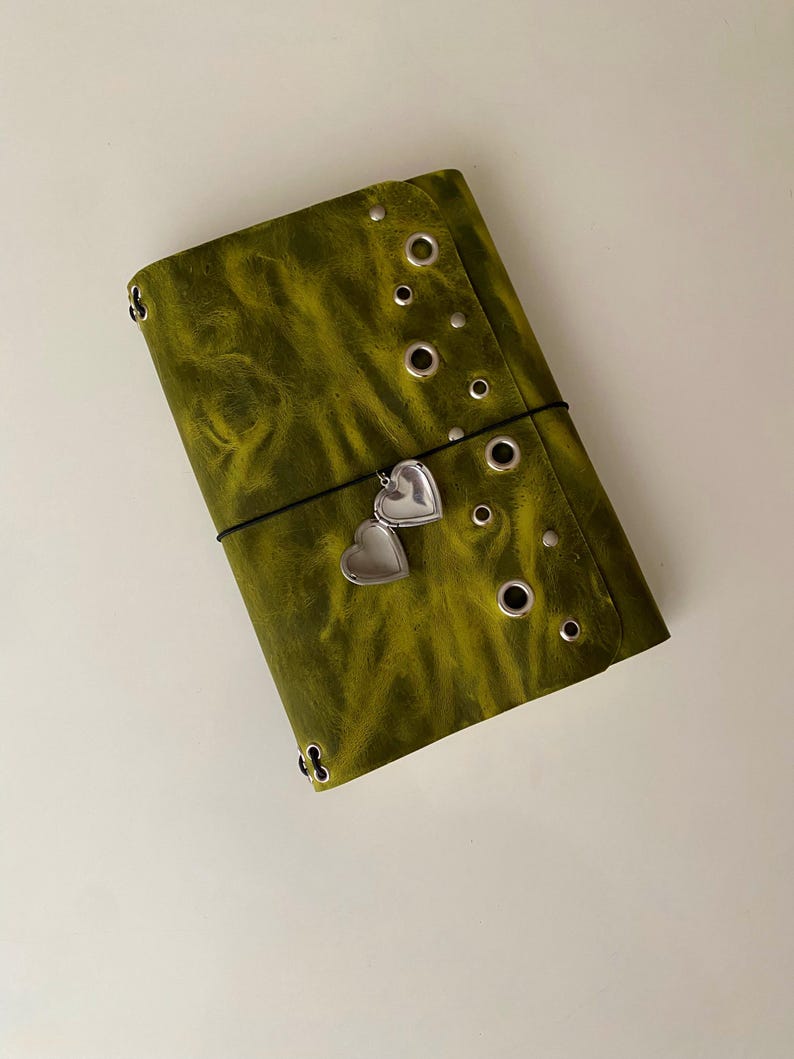 May include: A rectangular olive green journal cover with a distressed texture, featuring a vertical row of silver metal eyelets and studs along the right side. A thin black elastic band wraps around the center, holding a small silver heart-shaped locket.
