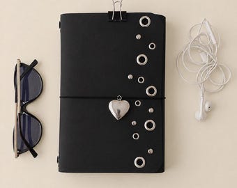 May include: A black notebook with a silver heart charm, metal eyelets, and a binder clip at the top. To the left are black-framed sunglasses, and to the right are white wired earbuds. The notebook is secured with a thin black elastic band.