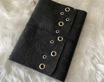 May include: A black journal cover featuring an embossed floral pattern across the surface. The front flap is secured with a series of silver-toned metal grommets arranged in a vertical line. A small silver star-shaped charm is attached to the top.