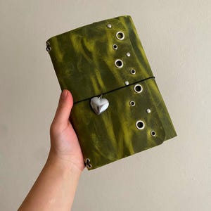 May include: A rectangular olive green leather journal cover featuring a distressed texture, multiple silver-toned metal grommets arranged along the right edge, and a black elastic cord closure adorned with a small, polished silver heart charm.