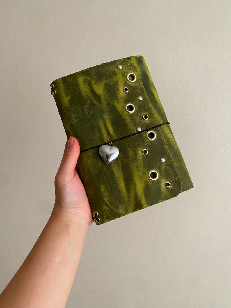 May include: A rectangular olive green leather journal cover featuring a distressed texture, multiple silver-toned metal grommets arranged along the right edge, and a black elastic cord closure adorned with a small, polished silver heart charm.