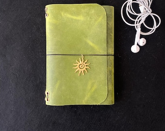 May include: A lime green leather journal with a wrap-around cover secured by a black elastic band. A gold-toned sun charm hangs from the center of the band. Beside the journal is a pair of white wired earbuds with a standard connector plug.