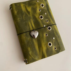 May include: A rectangular olive green journal with a distressed texture, featuring a vertical row of silver metal grommets and studs along the right edge. A thin black elastic band wraps around the center, holding a silver heart-shaped charm.