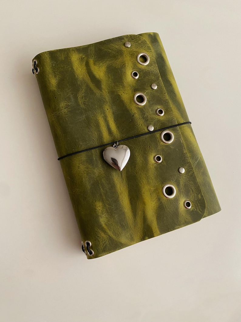 May include: A rectangular olive green journal with a distressed texture, featuring a vertical row of silver metal grommets and studs along the right edge. A thin black elastic band wraps around the center, holding a silver heart-shaped charm.