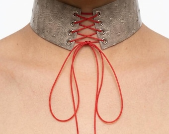 May include: A wide, textured metallic silver choker necklace featuring a front-facing corset-style lace-up design. The piece is secured with thin, vibrant red cord threaded through silver-toned metal eyelets, tied in a bow with long hanging ends.