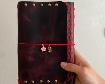 May include: A dark burgundy leather journal featuring a red elastic band closure, brass rivets along the top and bottom edges, and a red lace trim on the side. Two small charms, a pink flower and a gold letter S, hang from the elastic band.