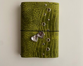 May include: A rectangular journal with a textured olive green crocodile-patterned cover. The cover features a vertical row of silver metal grommets and studs, secured by a thin black elastic band holding a silver heart-shaped locket charm.