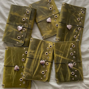 May include: Six rectangular olive green leather journals arranged in a group. Each journal features a distressed texture, a series of metallic eyelets along the side, and a thin black elastic band secured with a small, polished heart-shaped charm.