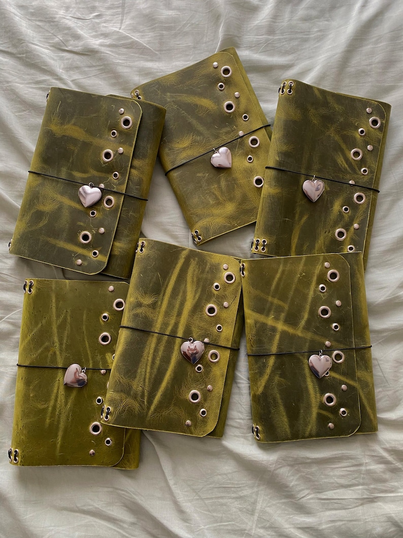May include: Six rectangular olive green leather journals arranged in a group. Each journal features a distressed texture, a series of metallic eyelets along the side, and a thin black elastic band secured with a small, polished heart-shaped charm.