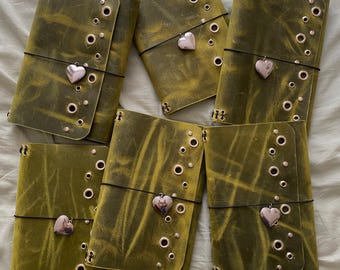May include: Five rectangular olive green leather journals arranged in a cluster. Each journal features a distressed texture, a series of silver-toned metal eyelets along the side, and a black elastic band secured with a silver heart-shaped charm.