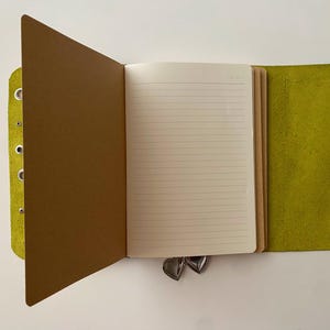 May include: A lime green leather journal cover with silver metal eyelets along the left edge. The open notebook features cream-colored lined paper pages and a brown paper divider. Two small silver heart-shaped charms hang from the bottom edge.