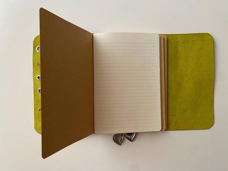 May include: A lime green leather journal cover with silver metal eyelets along the left edge. The open notebook features cream-colored lined paper pages and a brown paper divider. Two small silver heart-shaped charms hang from the bottom edge.