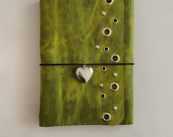 May include: A lime green leather journal cover featuring a vertical row of silver-toned metal grommets and studs along the right edge. A thin black elastic cord wraps horizontally around the cover, holding a small, polished silver heart-shaped charm.