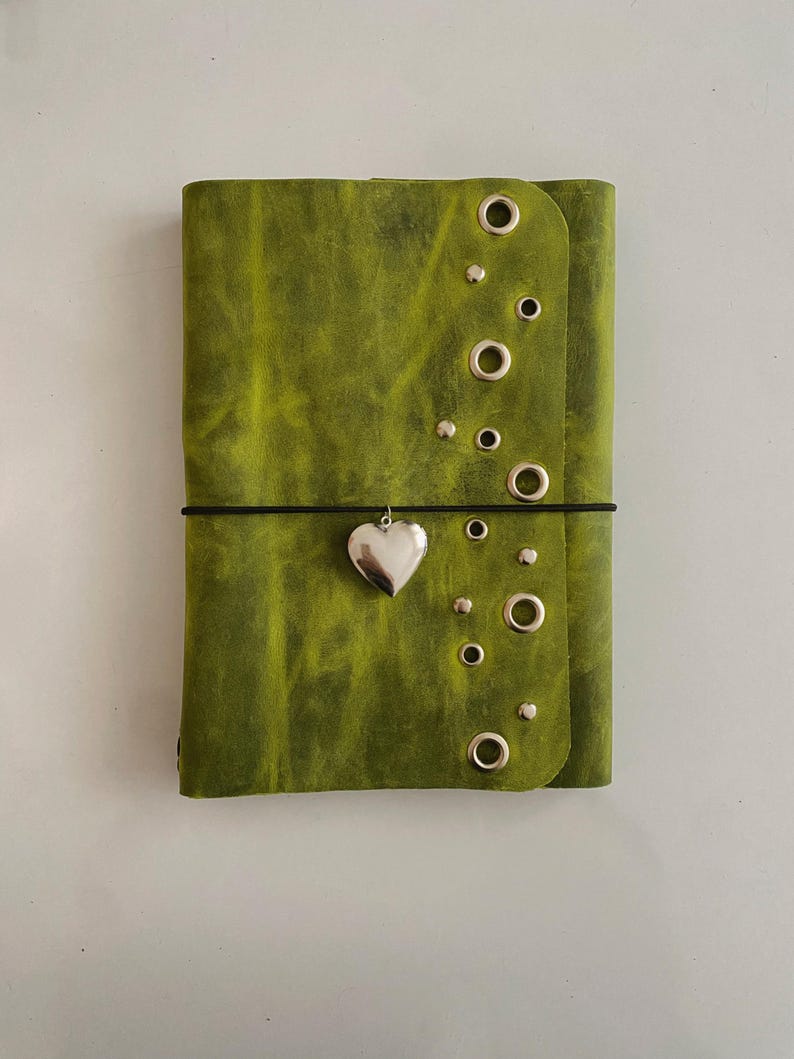 May include: A lime green leather journal cover featuring a vertical row of silver-toned metal grommets and studs along the right edge. A thin black elastic cord wraps horizontally around the center, holding a silver heart-shaped charm.