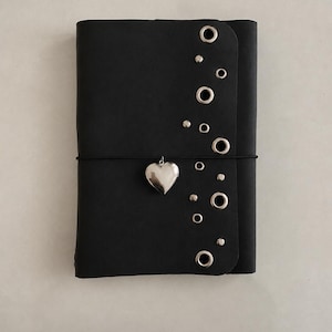 May include: A black notebook featuring a vertical row of silver-toned metal eyelets and small studs along the right edge. A thin black elastic band wraps horizontally around the center, holding a polished silver-toned heart-shaped charm.