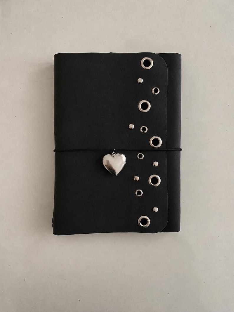 May include: A black notebook featuring a vertical row of silver-toned metal eyelets and small studs along the right edge. A thin black elastic band wraps horizontally around the center, holding a polished silver-toned heart-shaped charm.