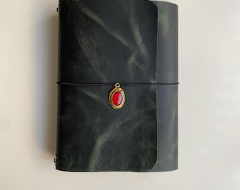 May include: A dark green leather journal with a wrap-around black elastic cord closure. The cord features a central gold-toned oval charm with a smooth, vibrant red stone inlay. The leather cover has a visible grain and subtle color variations.
