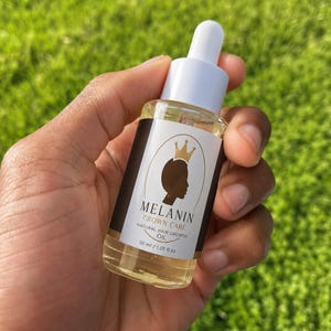 May include: A clear glass dropper bottle containing light yellow hair oil. The white label features a black silhouette of a person wearing a crown, with the text MELANIN CROWN CARE NATURAL HAIR GROWTH OIL and the volume 30 ml / 1.01 fl oz.