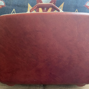 May include: A rectangular, deep red suitcase with a textured, leather-like finish. The suitcase features a sturdy, matching red top handle with silver-toned metal hardware attachments and rounded corners on the main body.