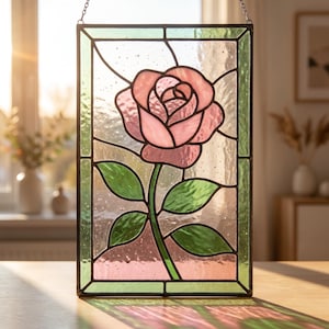 May include: A rectangular stained glass panel featuring a central pink rose with green leaves. The design is composed of textured glass pieces held together by dark metal lead lines. The panel is framed in a green border and hangs vertically.