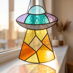 May include: A stained glass suncatcher shaped like a UFO, featuring a light blue dome, a teal and purple upper section, and a lower beam composed of yellow, orange, and amber glass panels. The piece is suspended by a thin metal chain.