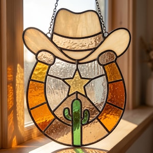 May include: A stained glass suncatcher featuring a beige cowboy hat positioned above a horseshoe shape. The horseshoe contains a central yellow star and a green cactus. The piece is constructed with dark metal lead lines and a hanging chain.