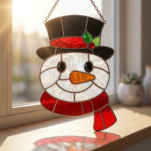 May include: A stained glass snowman suncatcher featuring a white textured face, a black top hat with a red band and green holly leaves, a bright orange carrot nose, black eyes, and a red scarf, all suspended by a metal chain for window display.