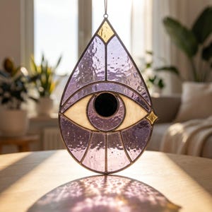 May include: A teardrop-shaped stained glass suncatcher featuring a central eye design. The piece is constructed with textured lavender glass, a solid black circular pupil, and cream-colored sclera, all held together by thin, dark metal solder lines.