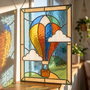 May include: A rectangular stained glass panel featuring a colorful hot air balloon in yellow, orange, and blue. The design includes white clouds against a textured blue sky, green rolling hills at the bottom, and a dark metal frame border.